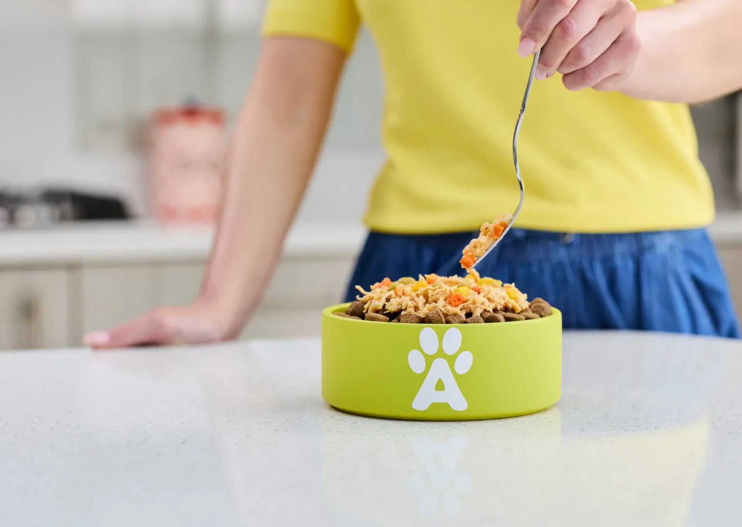 Woman in yellow top, blue jeans stirring a mixture of kibble and taste topppers in lime green Applaws bowl